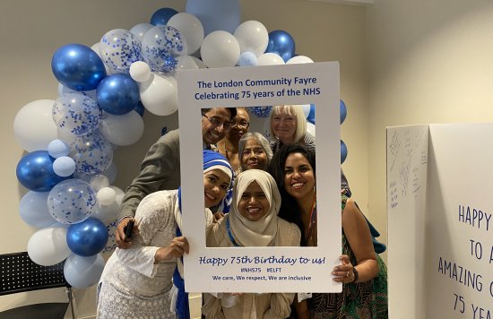 Photo of attendees at the East London Community Fayre, in front of a selfie frame, a balloon arch and a large birthday card.